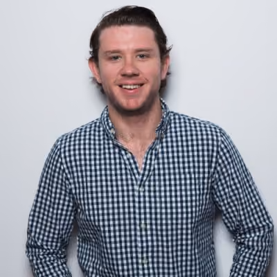 Smiling man with short brown hair wearing a blue and white checkered button-up shirt against a plain light background.