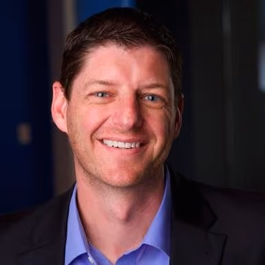 Smiling man with short brown hair wearing a black blazer and light blue shirt against a dark background.