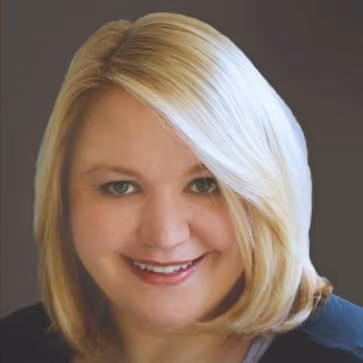 Portrait of a smiling woman with blonde hair and green eyes against a gray background.