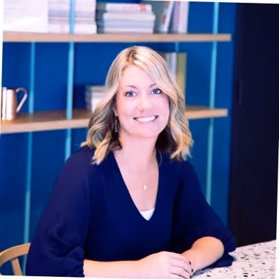 Smiling blonde woman with shoulder-length hair sitting at a table in front of a blue wall with shelves holding books and decor.