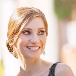 Smiling woman with light brown hair styled back, wearing black lace top and earrings, looking to the side.