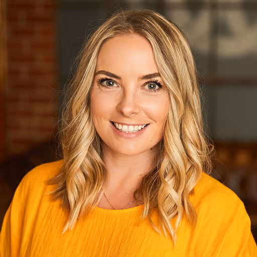 Smiling woman with blonde wavy hair wearing a bright yellow top against a blurred indoor background.