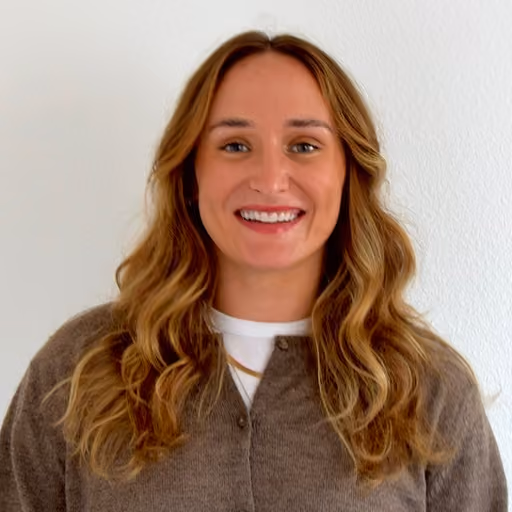 Smiling woman with long wavy blonde hair wearing a brown cardigan and white top against a plain white background.
