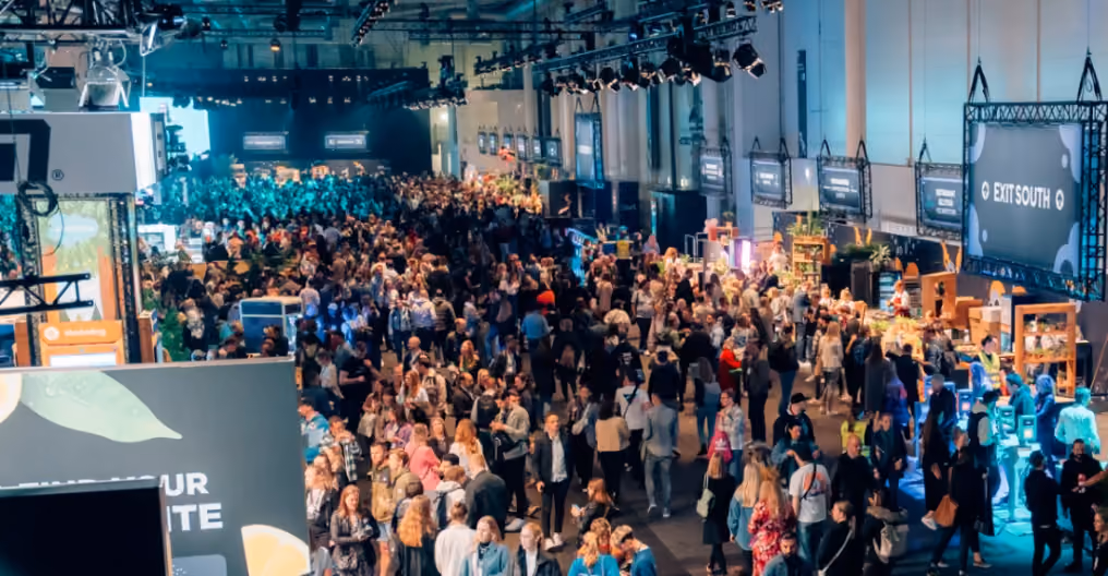 Large crowd of people attending an indoor event with various booths and a sign indicating 'EXIT SOUTH'.