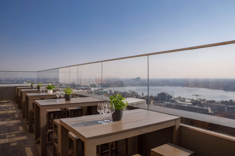 Rooftop terrace with tables set for wine, potted plants, and a panoramic city and river view under a clear sky.