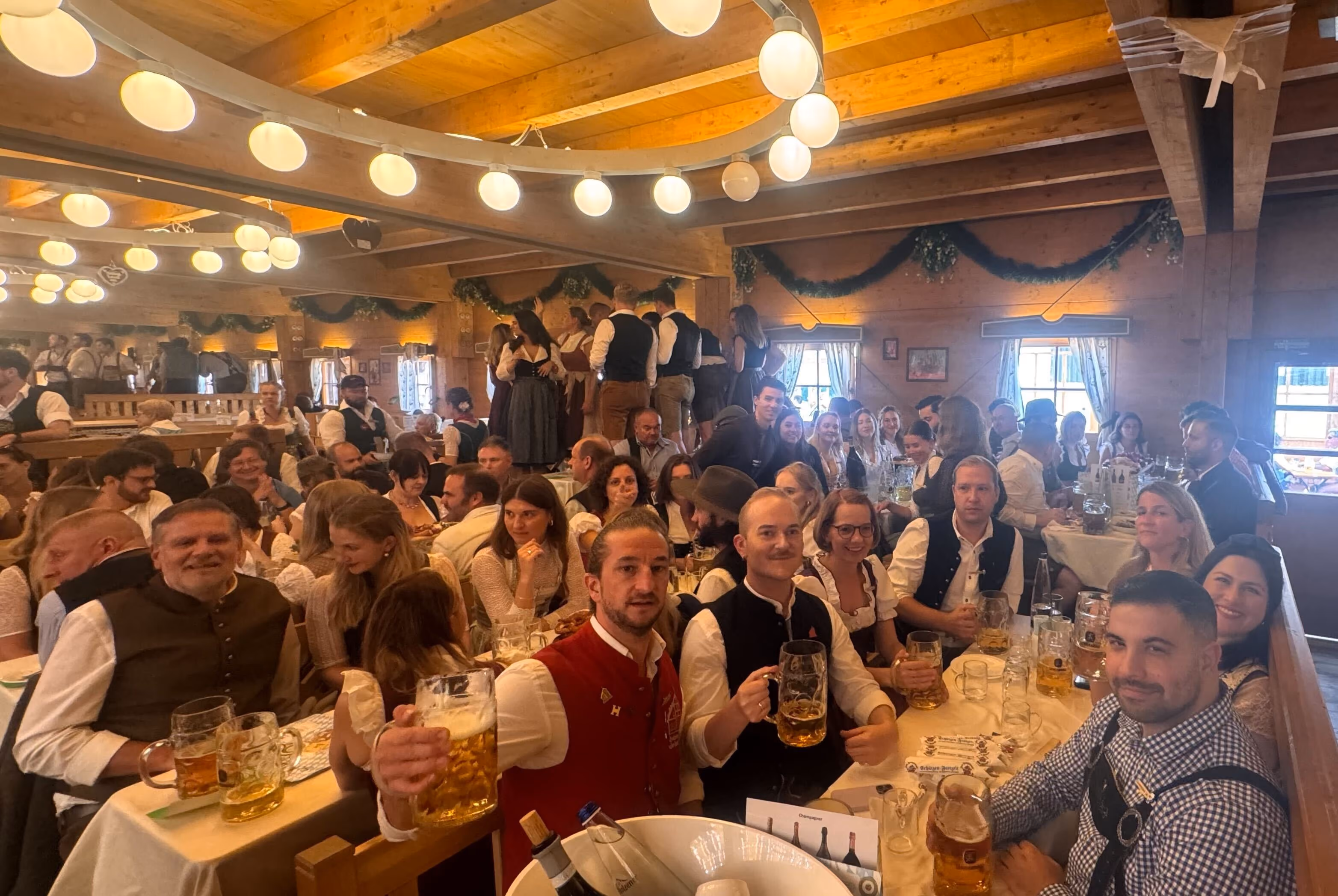 Crowded indoor Oktoberfest celebration with people in traditional Bavarian attire holding beer mugs at wooden tables under warm lighting.