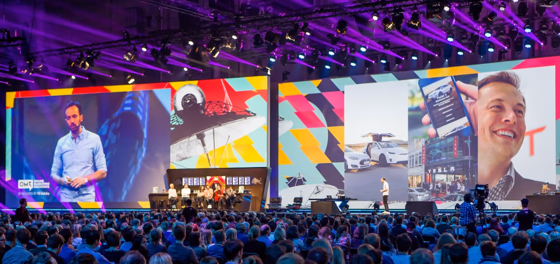 A large conference with an audience watching a speaker on stage with multiple big screens showing presentations, including images of Tesla cars and a smiling man.