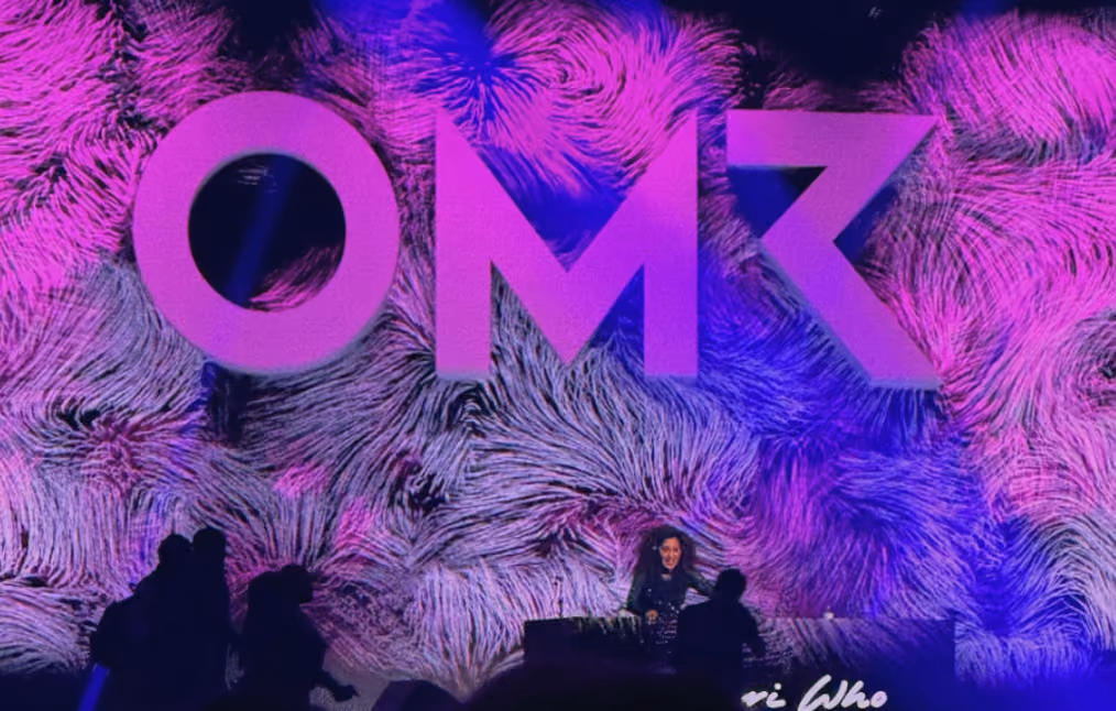 DJ performing in front of large purple OMR letters on a textured purple and blue backdrop with silhouetted audience in the foreground.