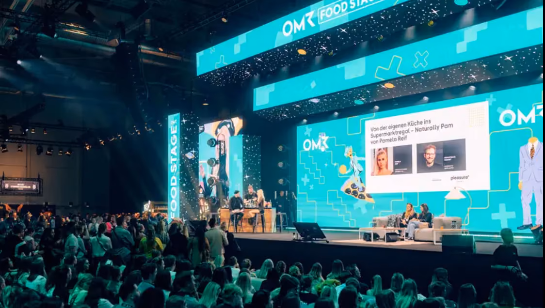 Large audience watching a panel discussion on the OM Food Stage with bright turquoise digital screens and speakers on stage.