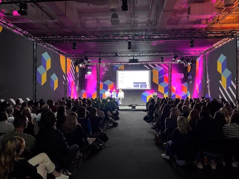 Audience seated facing a stage with three speakers and a large screen, with colorful geometric patterns and purple lighting on the walls.