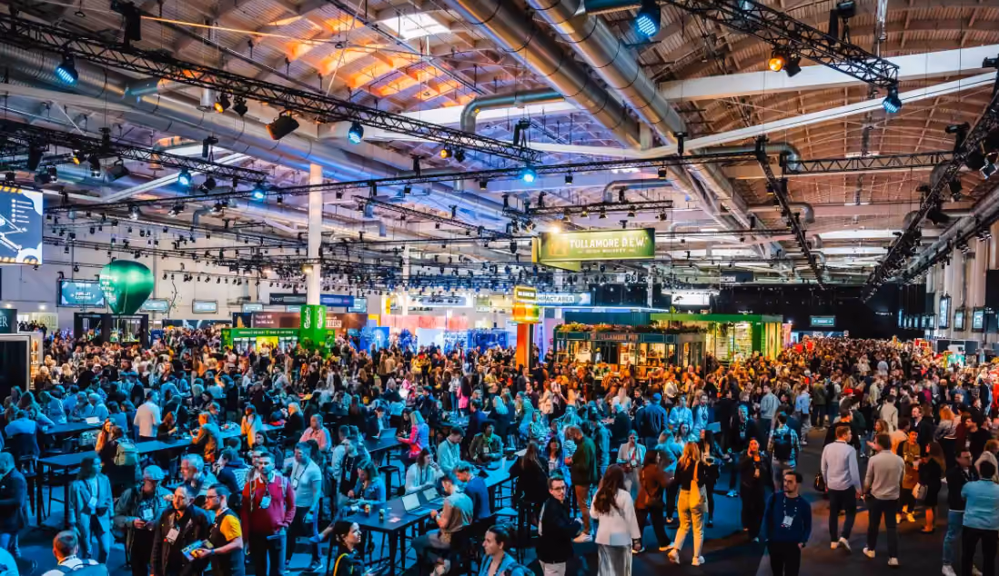 Crowded indoor convention or expo with many people walking and socializing under industrial ceiling lighting.