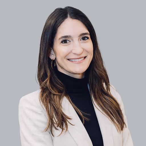 Smiling woman with long brown hair wearing a black turtleneck and beige blazer against a gray background.