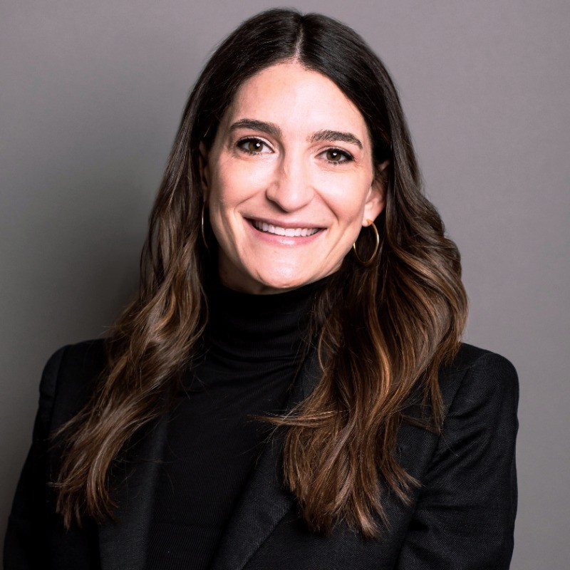 Smiling woman with long brown hair wearing a black turtleneck and beige blazer against a gray background.