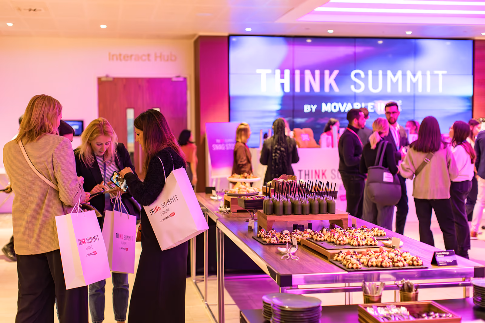 Attendees at THINK Summit Europe networking near a table with assorted snacks and drinks in a brightly lit conference area.