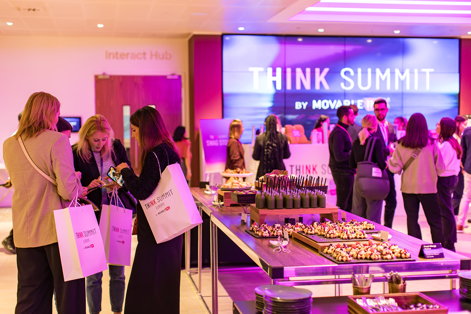Attendees at THINK Summit Europe networking near a table with assorted snacks and drinks in a brightly lit conference area.