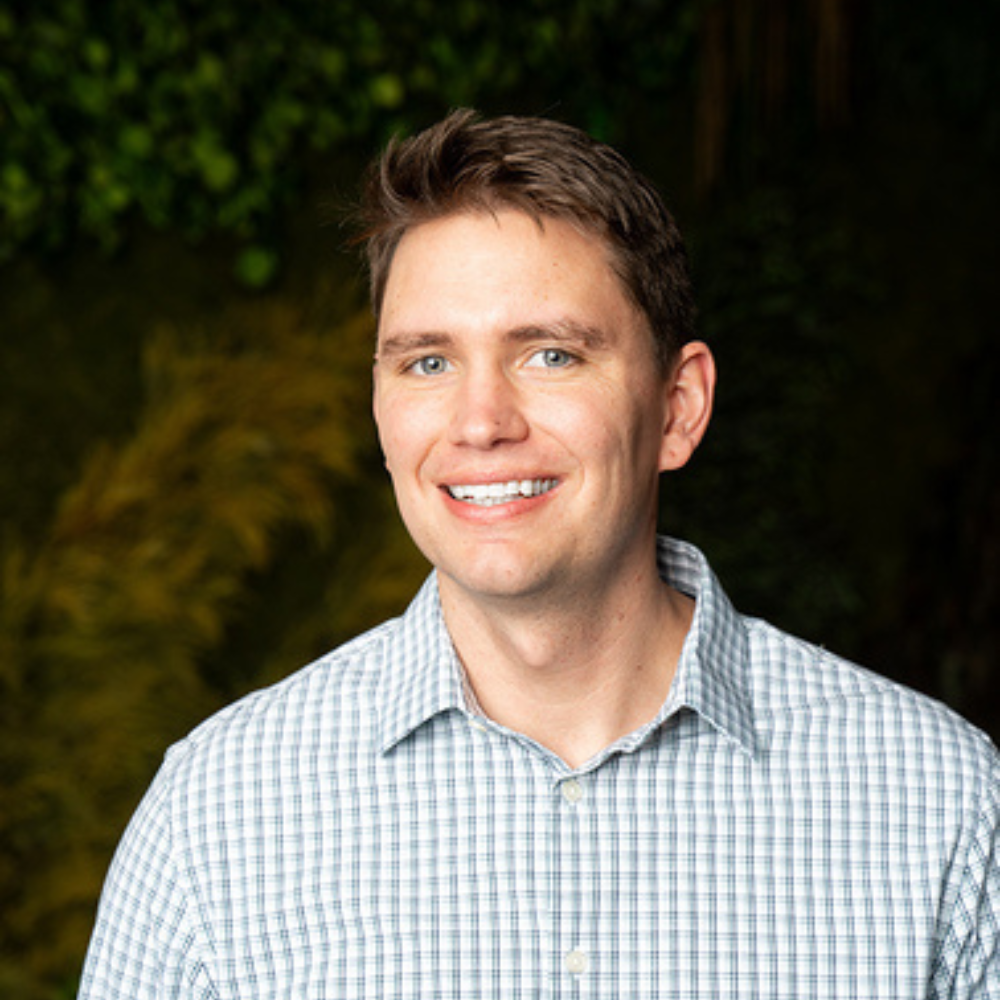 Smiling man with short brown hair wearing a light blue checkered button-up shirt against a blurred green and yellow foliage background.