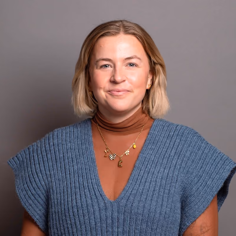 Smiling woman with short blonde hair wearing a brown turtleneck under a blue knit sleeveless sweater and a gold necklace with various charms.