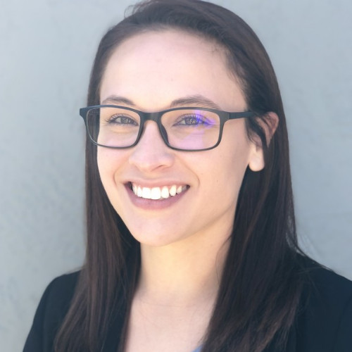 Smiling young woman with long dark hair wearing black glasses and a black blazer against a light gray background.