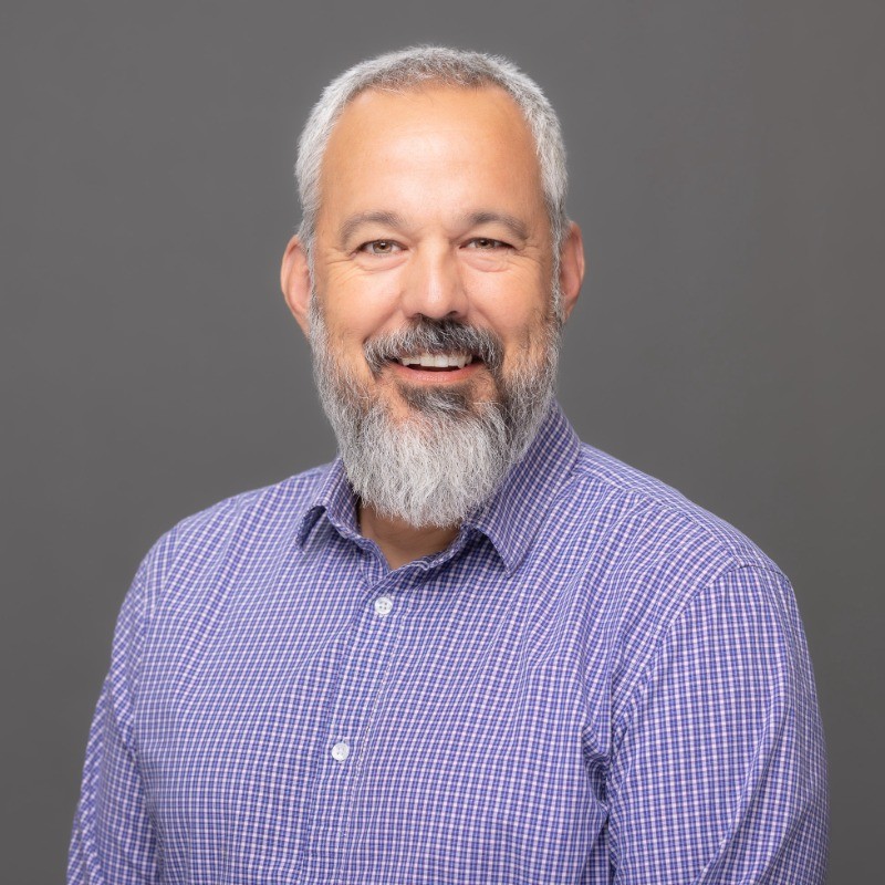 Smiling middle-aged man with gray hair and beard wearing a purple checkered shirt against a gray background.