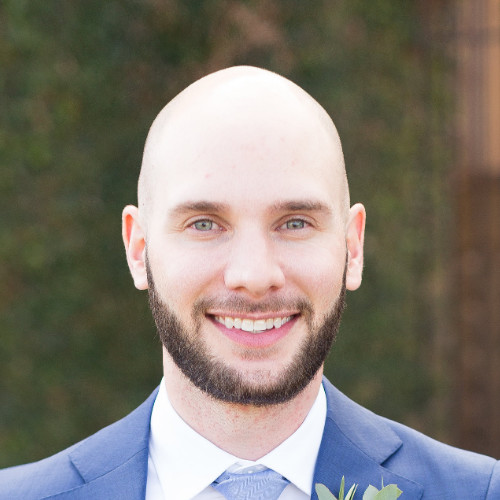Smiling bald man with a beard wearing a blue suit and light blue tie with a boutonniere.