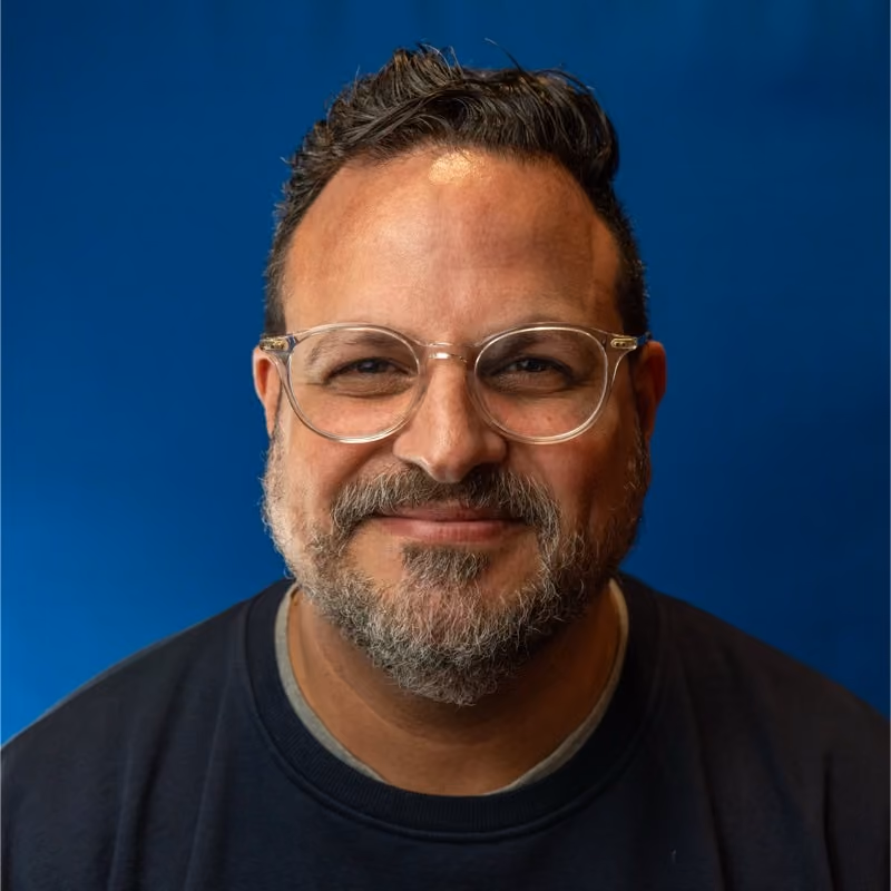 Smiling man with glasses, short curly hair, and a beard against a blue background.