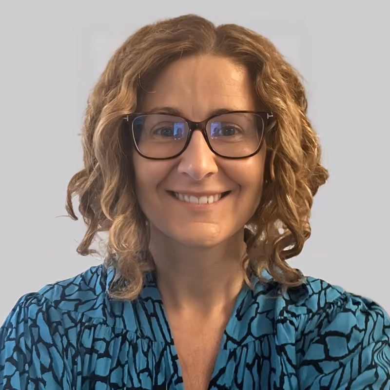 Smiling woman with curly light brown hair wearing dark rectangular glasses and a blue patterned blouse against a plain light background.