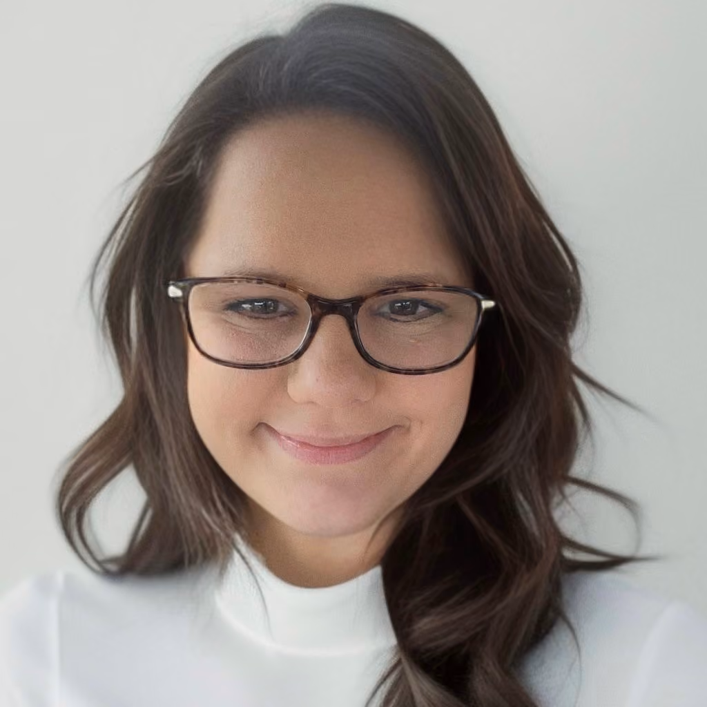 Woman with long dark hair and glasses smiling slightly against a plain light background.