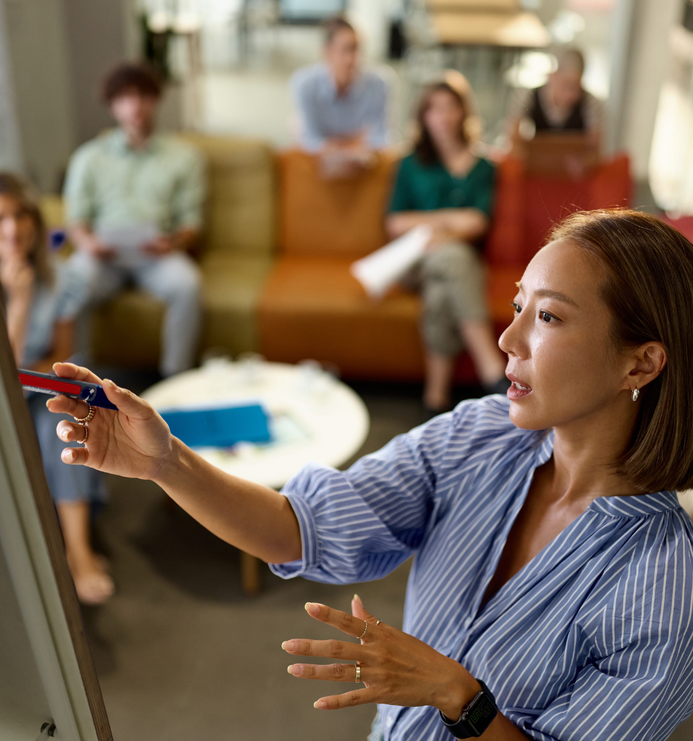 A woman in a blue and white striped shirt presenting and pointing at a whiteboard during a meeting, with five people sitting on colorful couches in the background attentively listening.