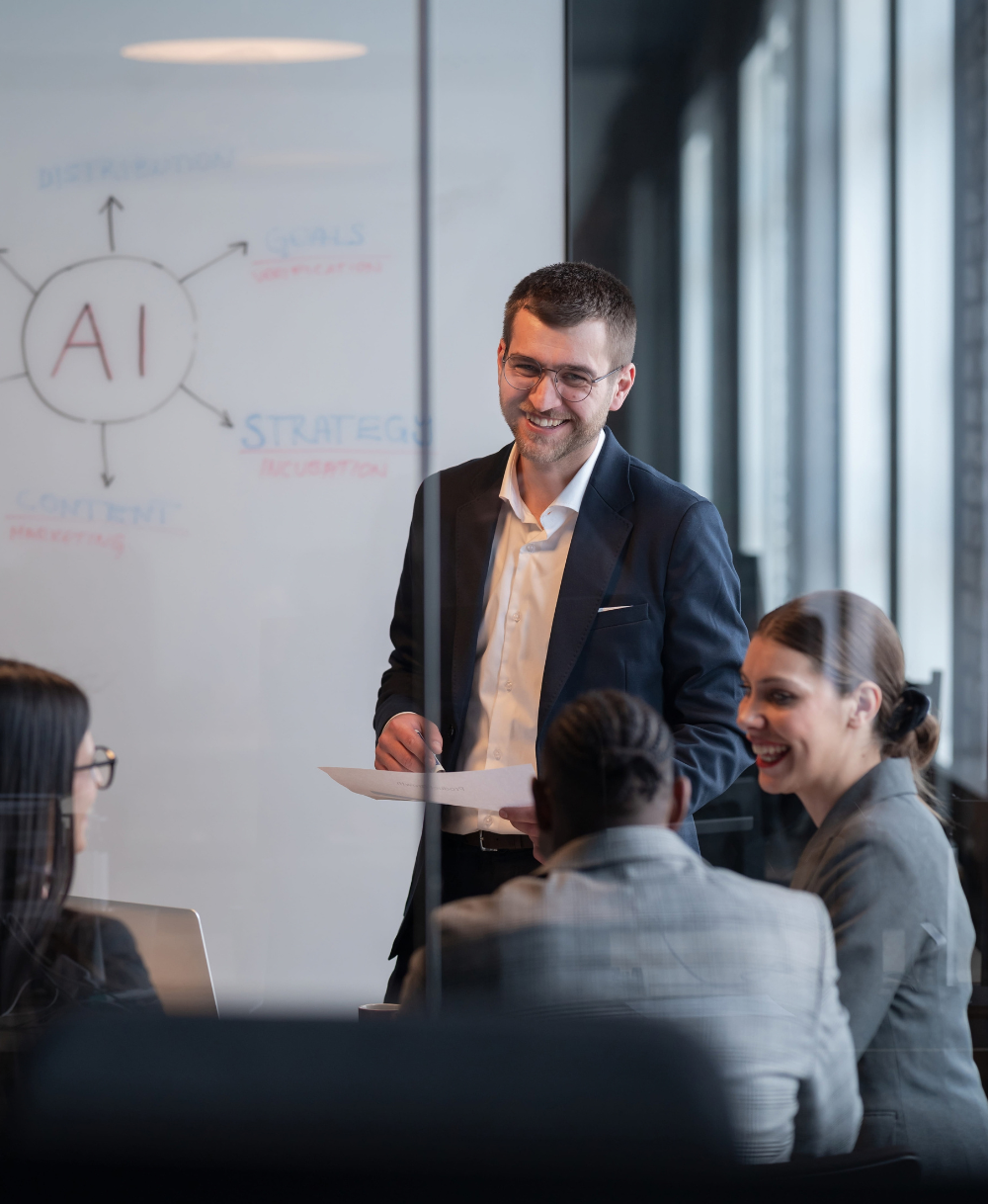 A man in a dark suit standing and smiling holds a paper while presenting to three seated colleagues in a glass-walled office with a whiteboard showing a diagram centered on 'AI' with related terms.