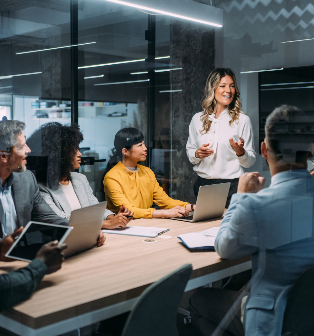 A diverse group of six business professionals having a meeting in a modern glass-walled conference room, with one woman standing and speaking while others listen attentively and use laptops and tablets.