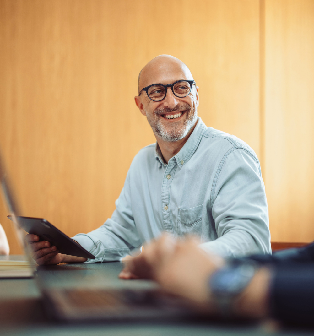 Smiling bald man with glasses holding a tablet, seated at a conference table with two other blurred people in a modern office setting.