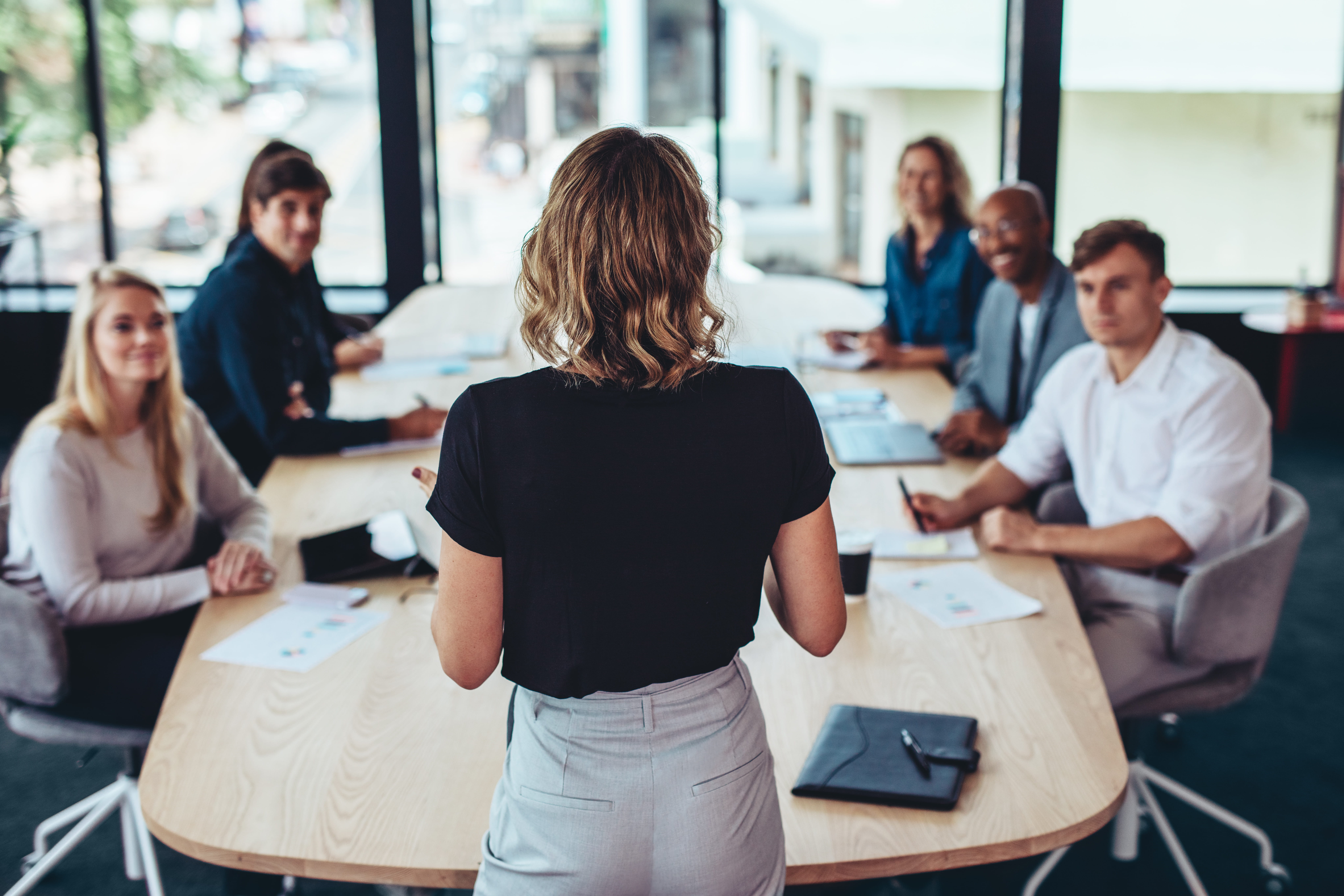 Woman standing at the head of a conference table speaking to five colleagues seated around a wooden table in a modern office with large windows.