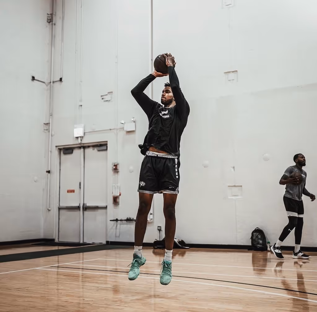 Basketball player in black sportswear jumping and shooting a basketball indoors on a wooden court.