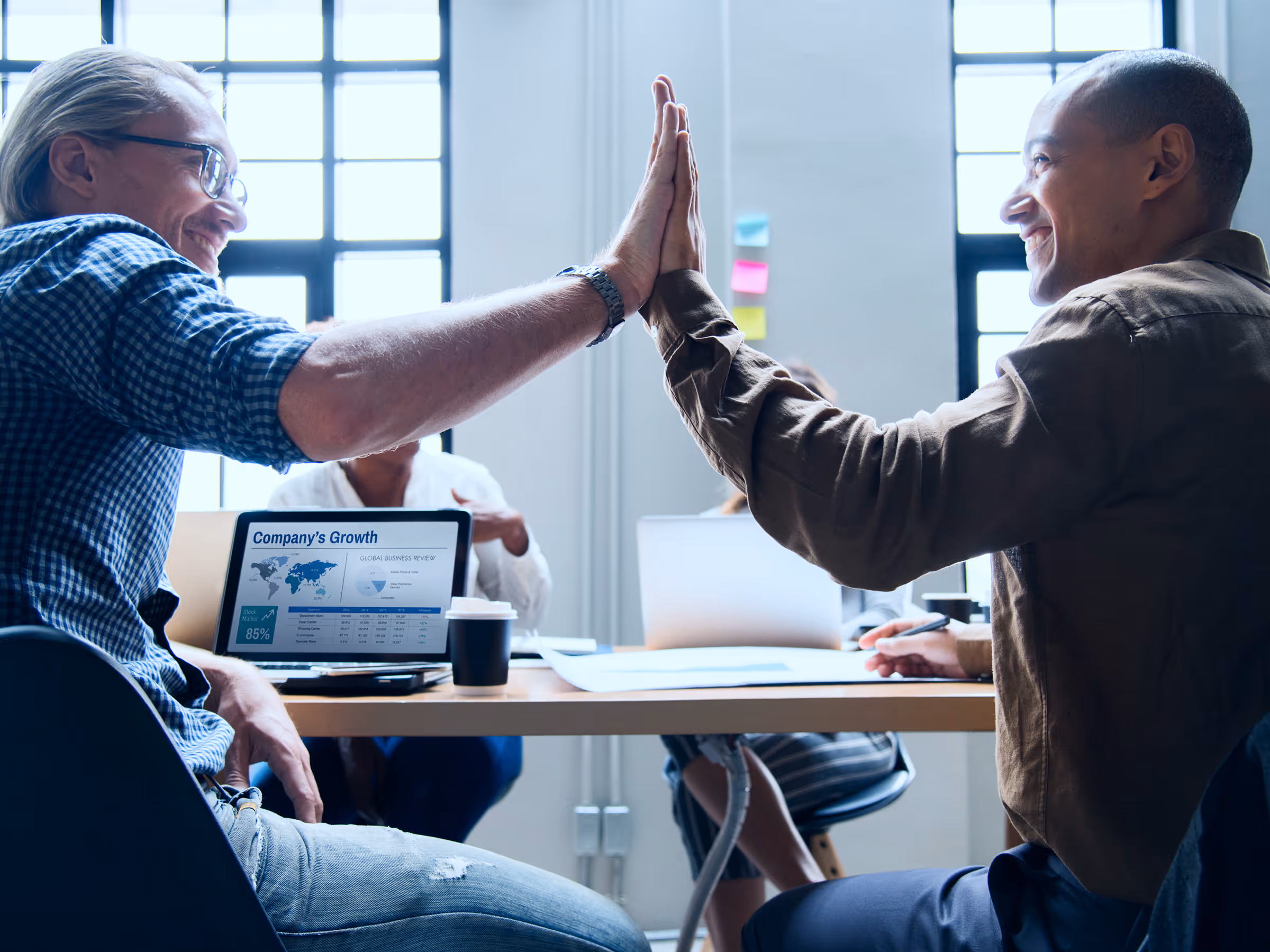 Two colleagues smiling and giving a high-five in a bright office with a laptop displaying company growth data on the desk.