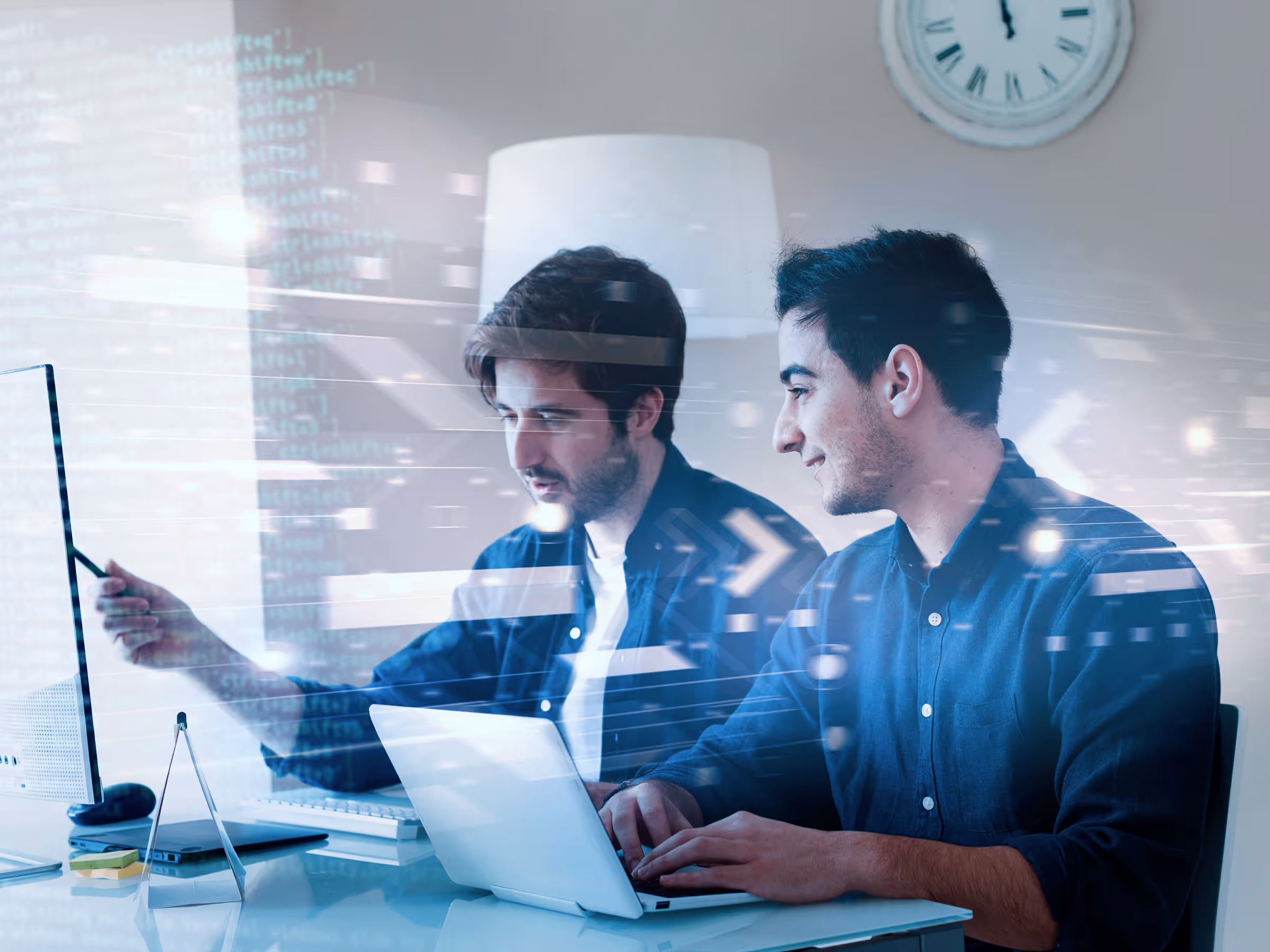 Two men collaborating at a desk, one pointing at a computer screen and the other typing on a laptop with digital data overlays.