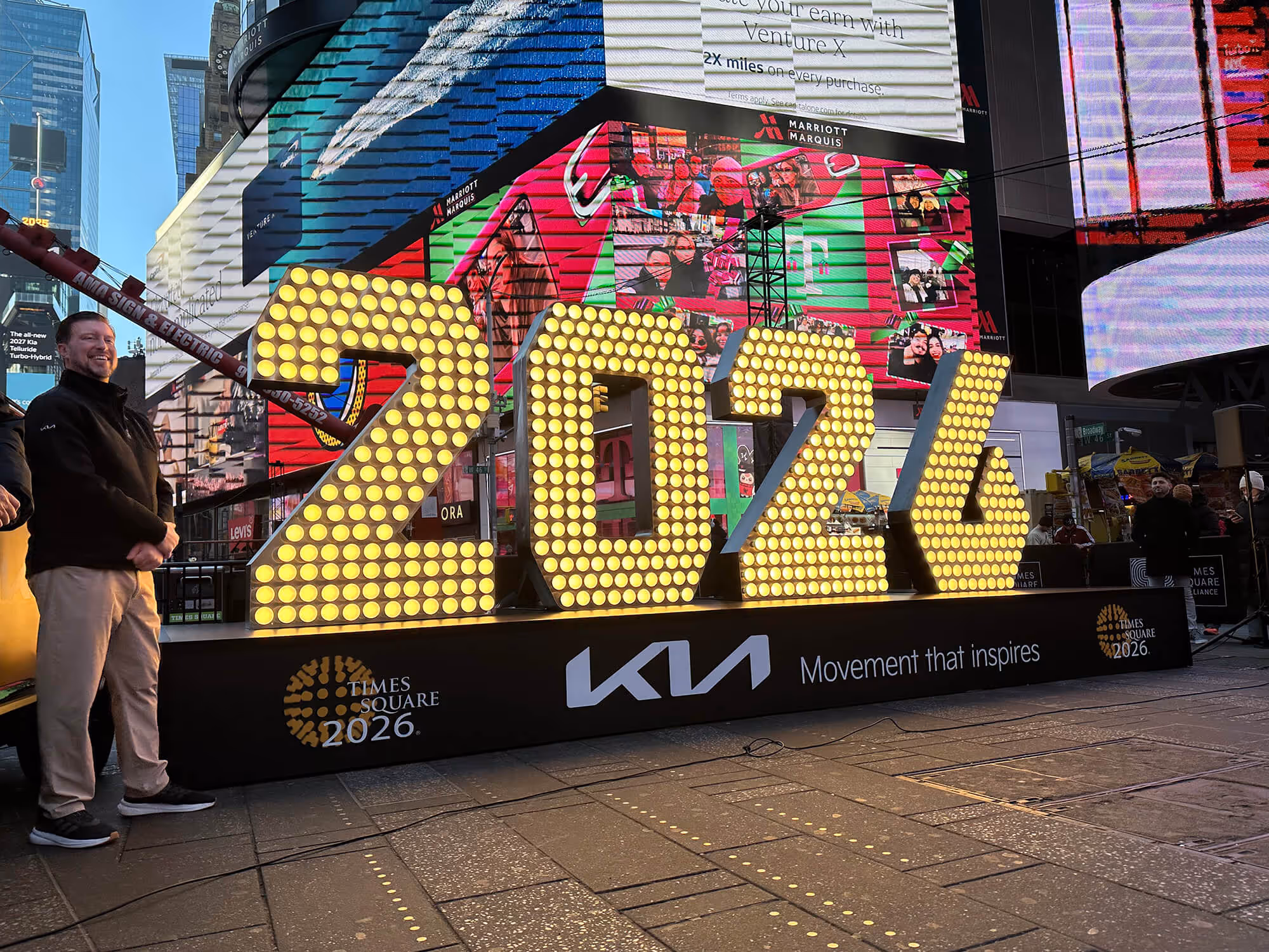 The numerals '2026,' each six feet tall and covered in lightbulbs, in Times Square on a platform with logos for KIA and Times Square New Year's Eve.