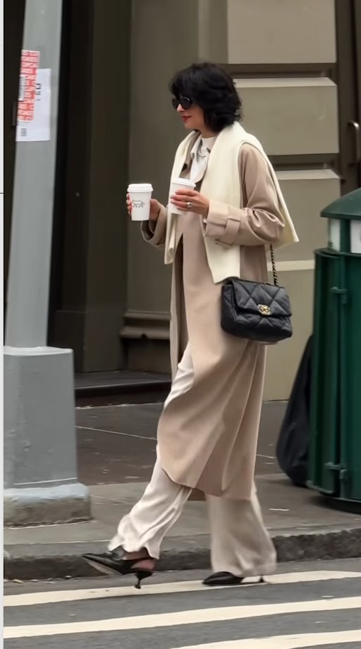 Influencer in a beige coat and sunglasses walking down a city street holding coffee