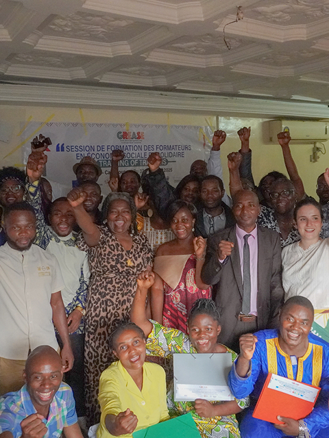 A group of people posing together with raised fists in front of a banner that reads "SESSION DE FORMATION DES FORMATEURS" and "TRAINING OF TRAINERS."