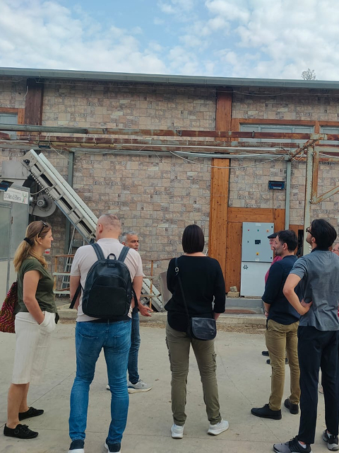 A small group of people standing outdoors, listening to a man giving a tour or explanation in front of a stone building with industrial machinery and a conveyor belt.