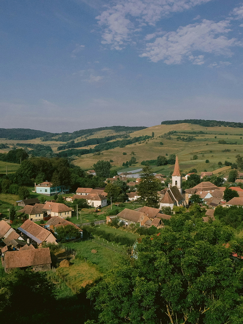  An elevated view of a small village with red-roofed houses and a prominent church steeple, nestled in a valley of rolling green hills under a clear sky.