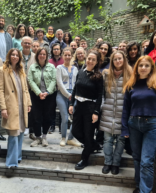  A large group of smiling colleagues or peers posing for a group photo on outdoor stone steps, with a brick wall and green climbing plants in the background.