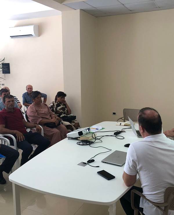  A man leading a presentation or meeting for a small group of people seated around a white table with laptops and a projector.