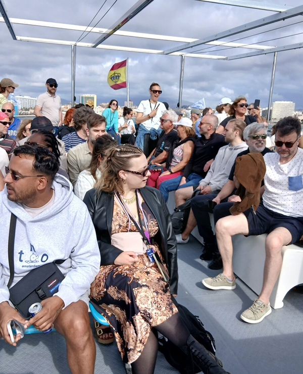  A large group of people sitting on the deck of a boat under a cloudy sky, with a Spanish flag flying in the background and a city coastline in the distance.