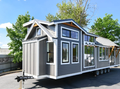 Exterior of a TruForm Tiny home on wheels with a white and dark wood facade, multiple tall windows, rooftop loft window, and propane setup on a dual-axle trailer parked outdoors.