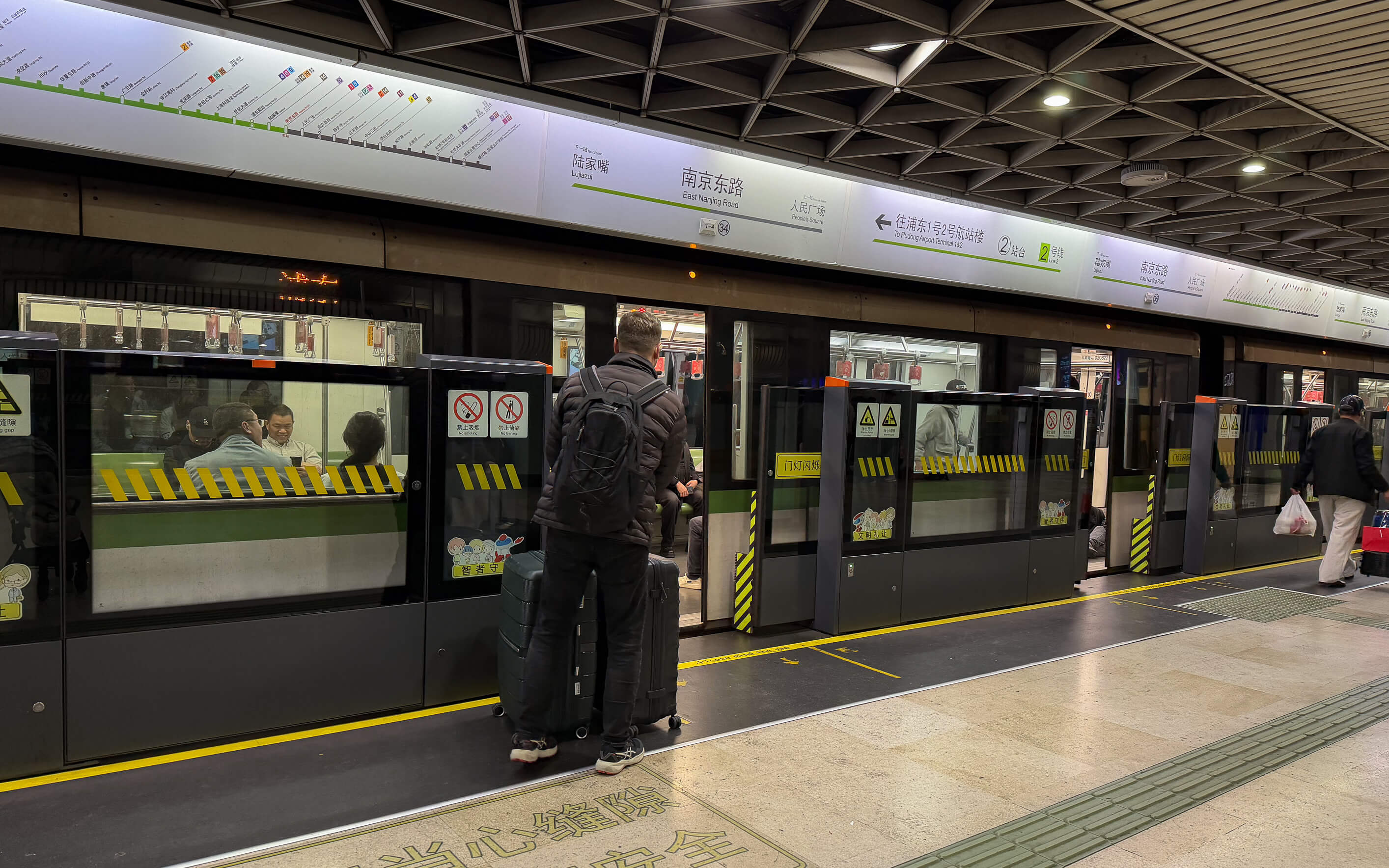 Chinesische Shanghai Metro Bahnsteig: Grüne U-Bahn mit Sicherheitsmarkierung, wartende Passagiere und Informationstafel in chinesischer Sprache – modernes Transportsystem