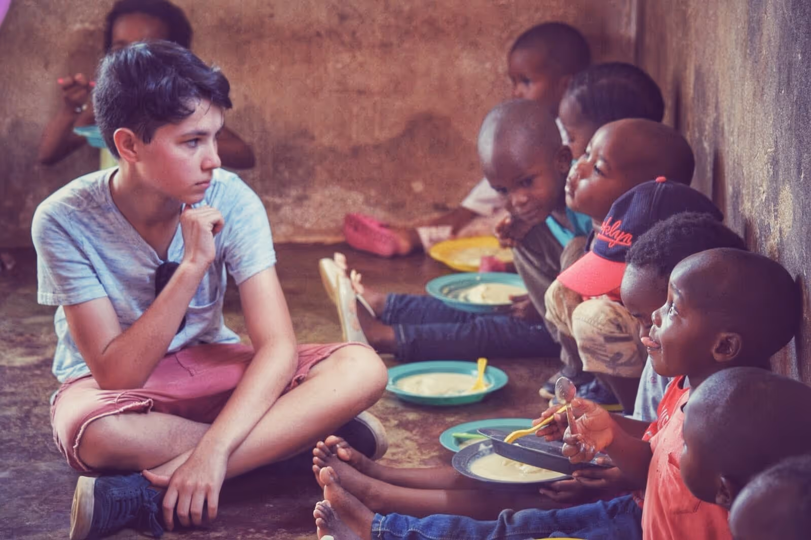 Nederlandse student zit op de grond en kijkt verwonderd naar de Zuid-Afrikaanse kinderen naast hem met een bord pap op hun schoot.