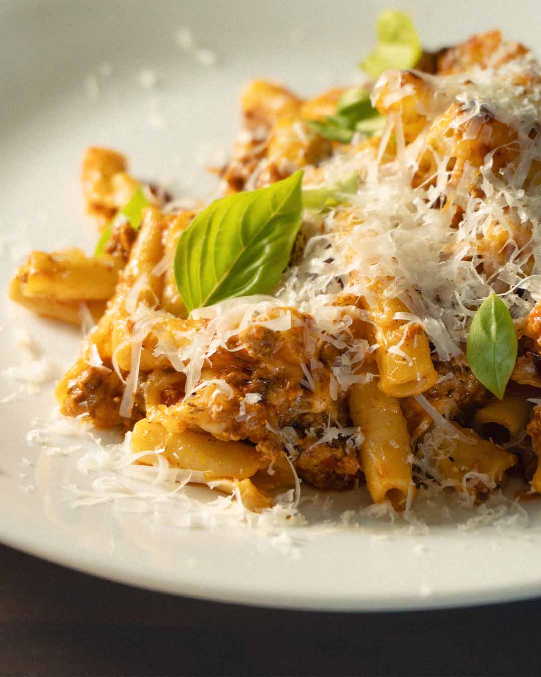 A close-up, atmospheric macro shot of a warm serving of baked ziti on a white plate, featuring a rich, dark meat sauce made with Mt. Folly ground beef, freshly grated parmesan cheese, and vibrant green basil leaves under moody, directional lighting.