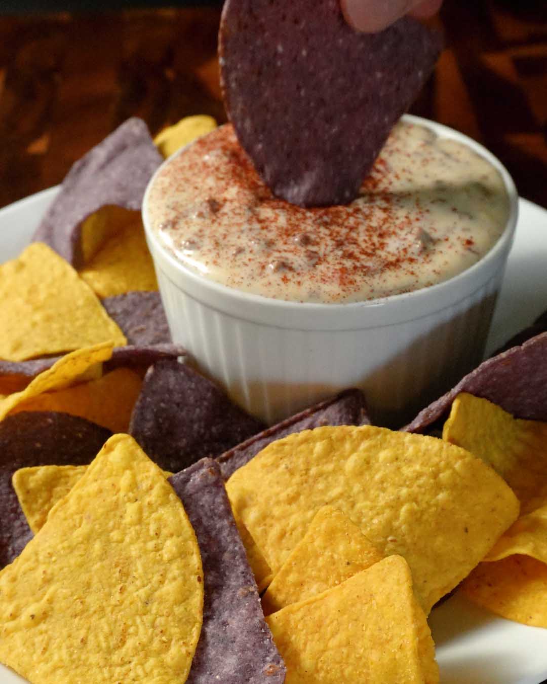 A blue corn tortilla chip dipping into a bowl of warm, creamy beef queso made with Mt. Folly pasture-raised ground beef and garnished with paprika.