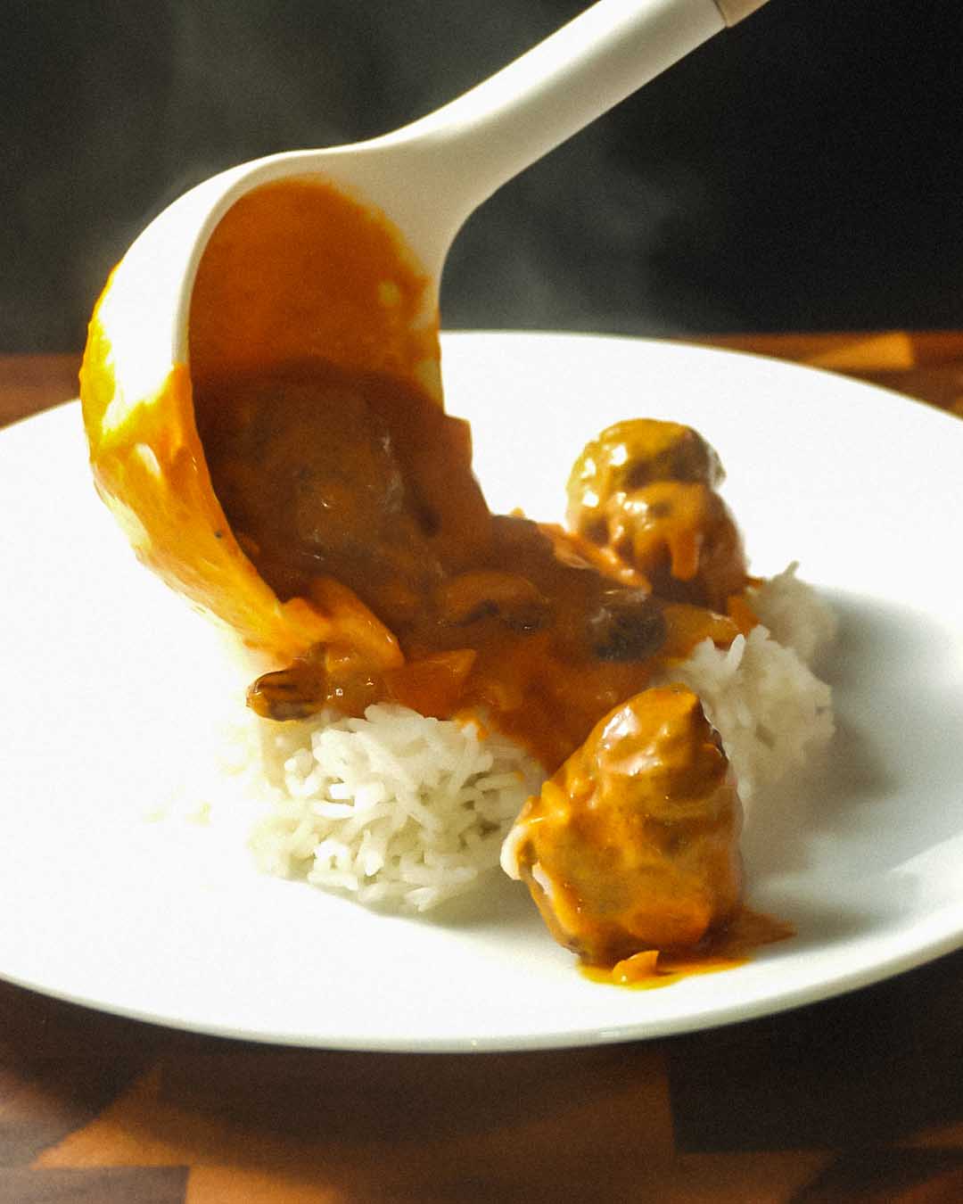 A white ladle pouring steaming Kentucky Kofta Masala—a rich ground beef curry—over a bed of fluffy basmati rice. The moody, dark background highlights the vibrant spiced sauce and Mt. Folly beef meatballs.