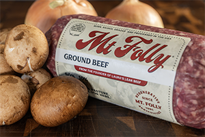 A 1-pound roll of pasture-raised Mt. Folly ground beef on a dark wooden cutting board, flanked by fresh brown cremini mushrooms and whole yellow onions.